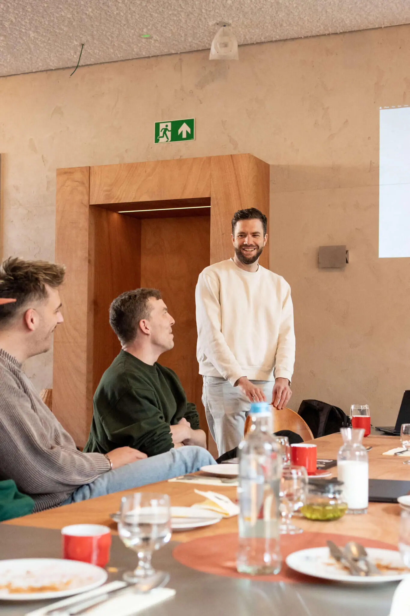 A smiling colleague in a cream sweater stands at a long lunch table while two others laugh