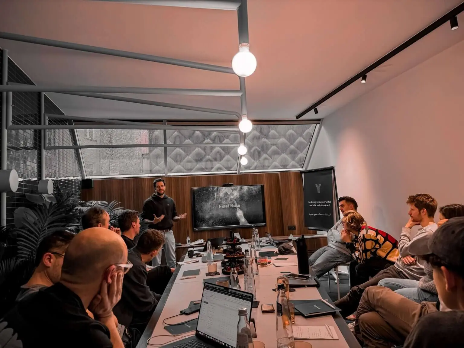 A colleague presents to the team around a long meeting table, the screen showing a slide titled Final Note