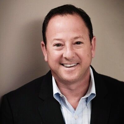 A man with short dark hair and wearing a dark suit jacket and light blue shirt smiles at the camera against a plain background, representing the warmth and professionalism associated with effective employee training.