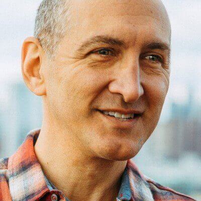 A man with short hair, wearing a checkered shirt, smiles while looking slightly to the right. The background is blurred, showing an outdoor setting, possibly an urban area. The scene evokes a sense of confidence and readiness akin to thorough employee training.