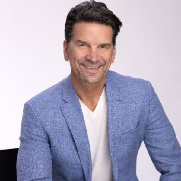 A man with short dark hair and a beard, wearing a light blue jacket and white shirt, smiles while seated against a plain background, possibly ready for an onboarding session.