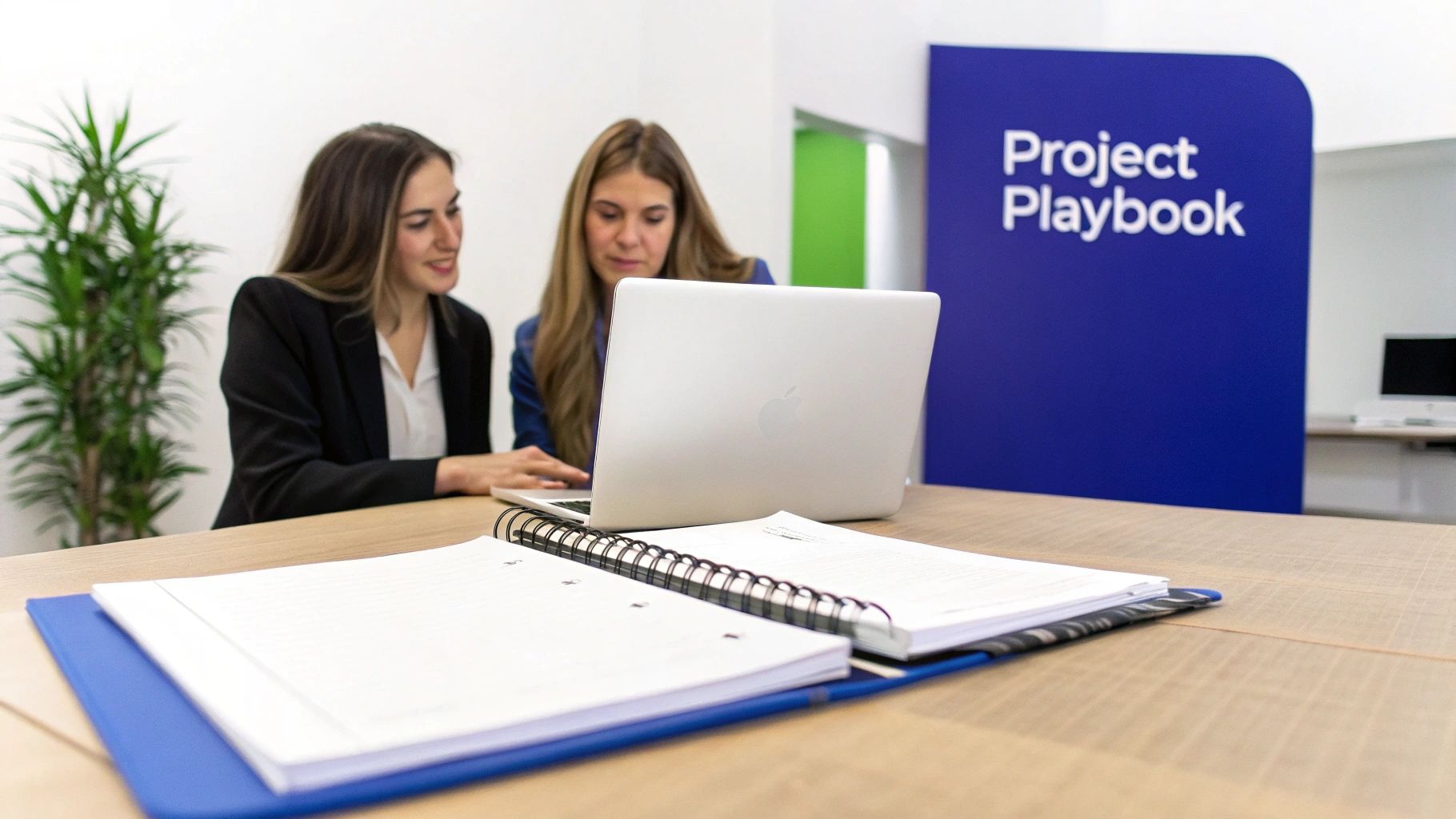 Two women collaborate on a laptop at a desk with notebooks, in an office setting with a 'Project Playbook' sign.