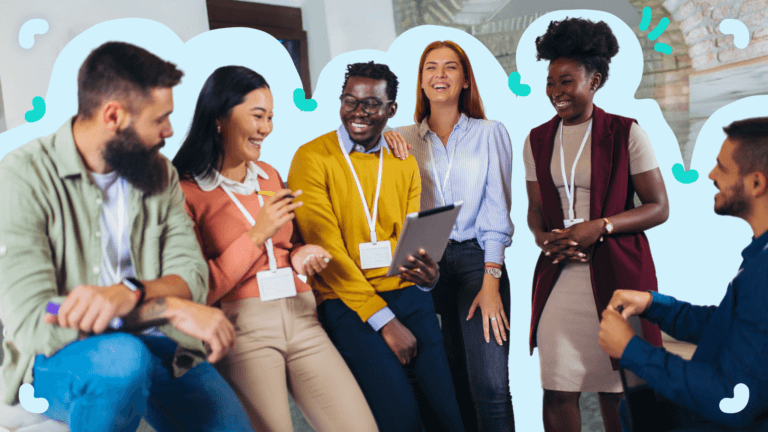 A group of six diverse people wearing conference badges are sitting and standing, smiling and engaging in a lively conversation about the new training matrix, with one holding a tablet.