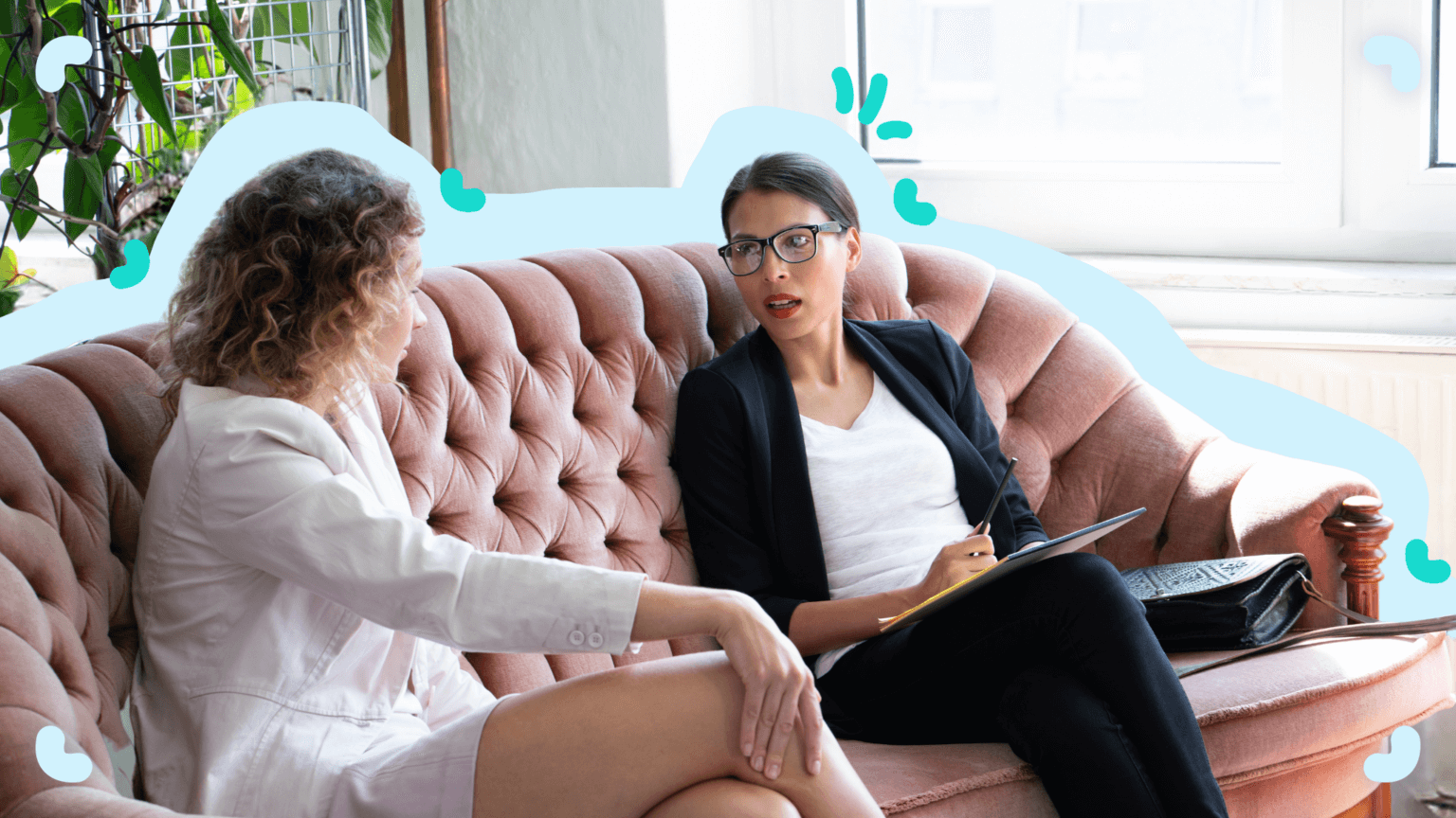 Two women, one in a white suit and the other in a black blazer, sit on a pink tufted sofa engaged in conversation. The woman in black holds a notebook and pen, discussing process documentation.