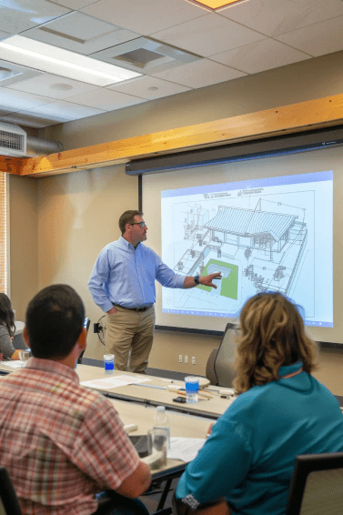 A man in a blue shirt stands in front of a projected architectural blueprint, pointing at the screen, discussing roofing details while several people seated at a table listen attentively in a meeting room.