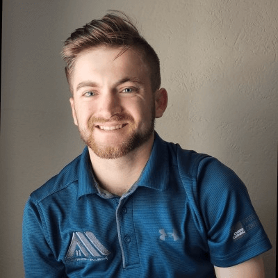 A man with short hair and a beard, wearing a blue collared shirt with industry logos, smiles while facing the camera.