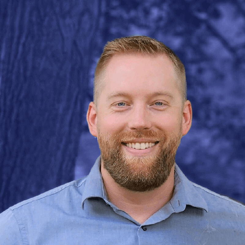 A man with a beard and short hair smiles at the camera, capturing a moment of positivity against a blurred blue and purple outdoor background. His confidence exudes the benefits of thorough employee training.