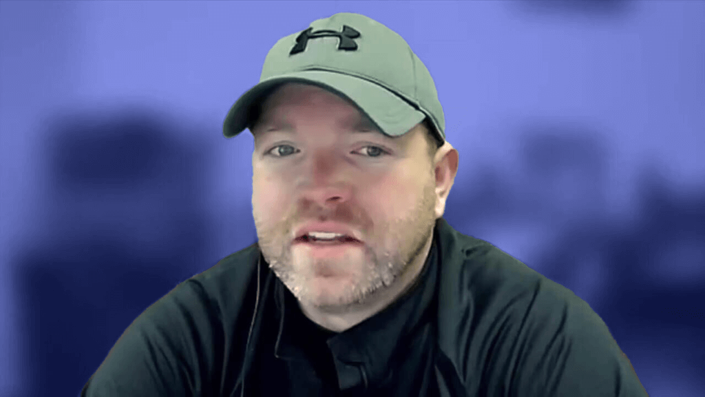 A man wearing a gray baseball cap and a dark shirt stands before a blurred blue backdrop, perhaps ready for an onboarding session.