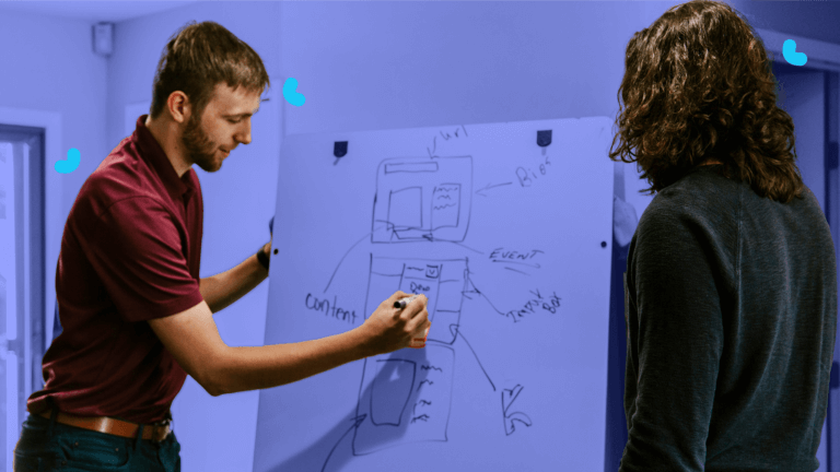 Two individuals stand next to a whiteboard covered in sketches and notes. Discussion on org chart examples