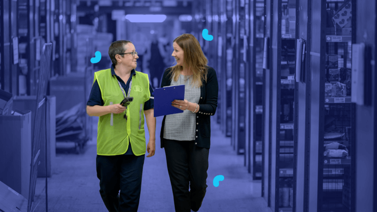 Two people are walking and talking in a warehouse aisle. One is wearing a high-visibility vest and holding a scanner; the other is holding a clipboard, discussing SOPs to ensure accurate process documentation.