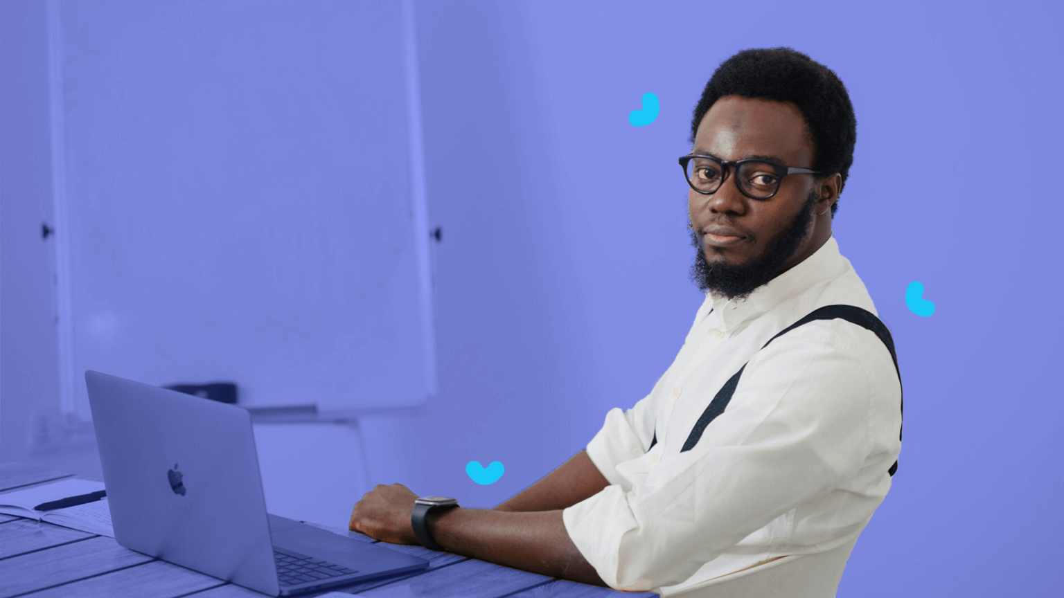 A man with glasses and a beard sits at a desk with a laptop, wearing a white shirt and suspenders. A whiteboard in the background outlines various roles and responsibilities.