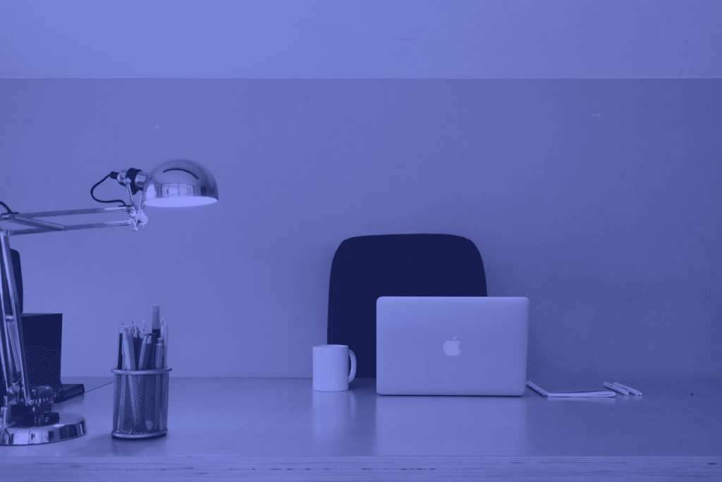 A minimalist workspace, ideal for employee training, features a laptop, a desk lamp, a mug, a pencil holder, and a notepad with pens, all arranged neatly on a desk with a chair. The image has a blue tint.