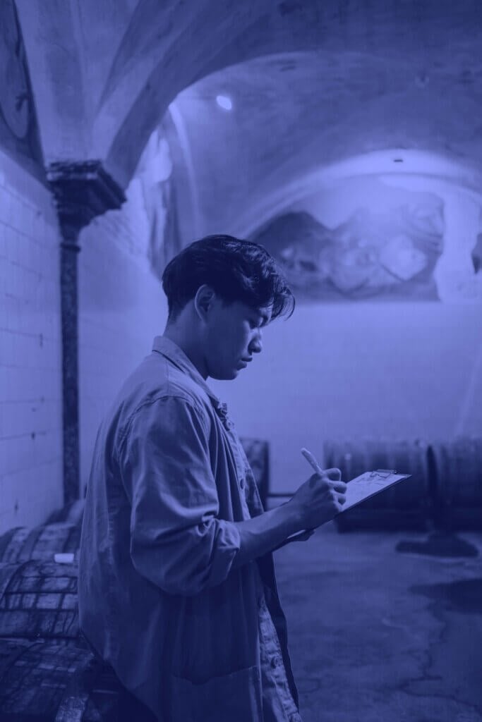 A person with short dark hair writes on a clipboard while standing in a dimly lit room with arched ceilings and large barrels in the background, seemingly focused on updating SOPs for employee training.