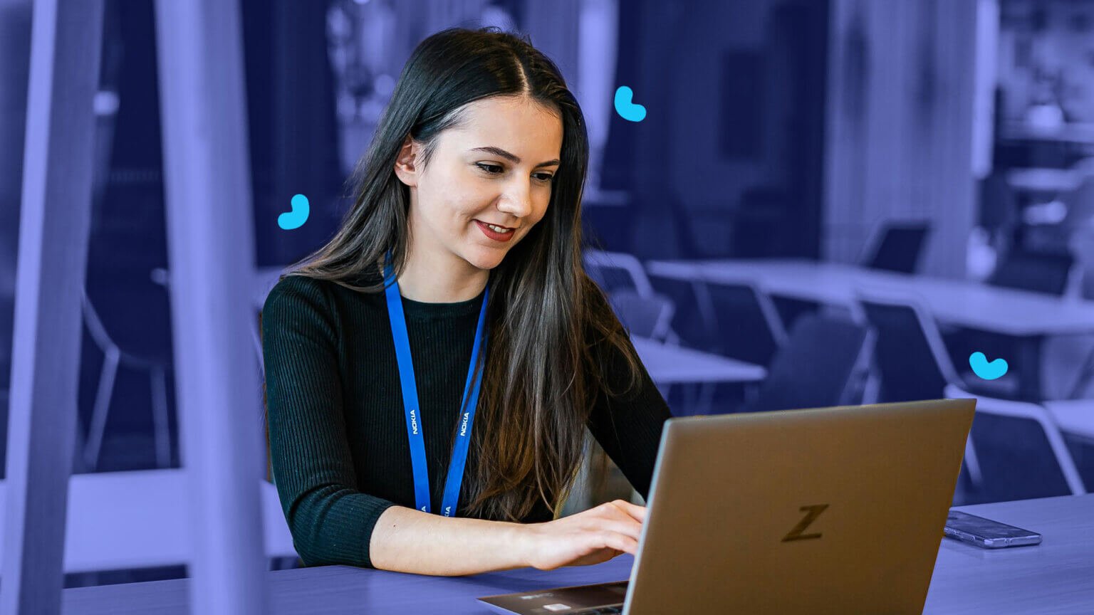 A woman with long dark hair, wearing a lanyard, is sitting at a desk in a modern office setting, engrossed in using her laptop to review AI training materials.