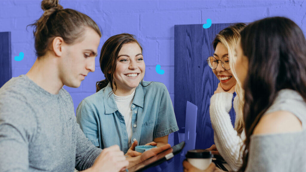 A group of four young adults, two men and two women, sit together at a table, smiling and talking. Each person has a beverage. The background features a blue wall adorned with posters about employee onboarding.